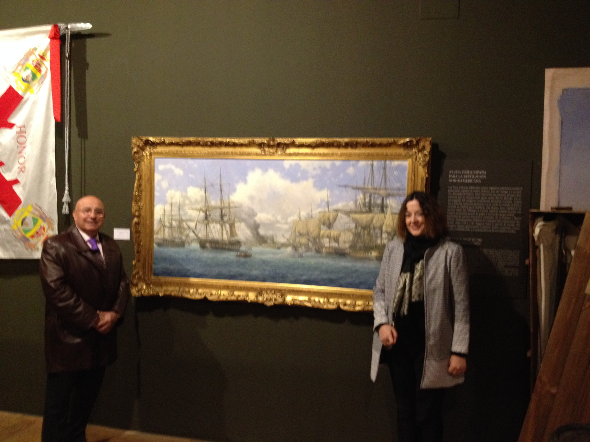 Dos visitantes junto al óleo de Pensacola en su marco dorado, colgado en la sala de Casa de América al lado de una bandera coronela