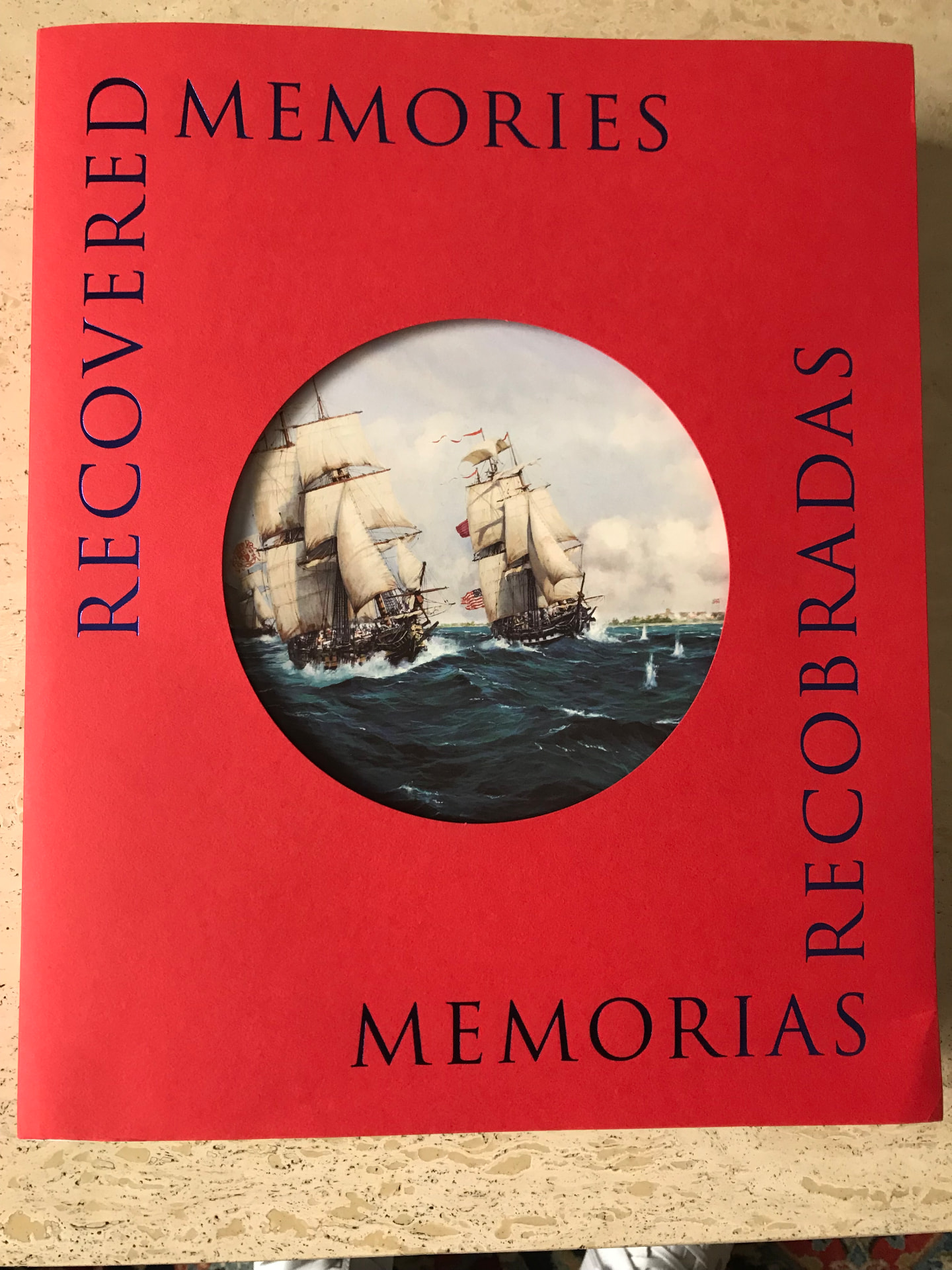 Cubierta roja del catálogo «Recovered Memories / Memorias recobradas» con una ventana circular troquelada que muestra dos fragatas en combate