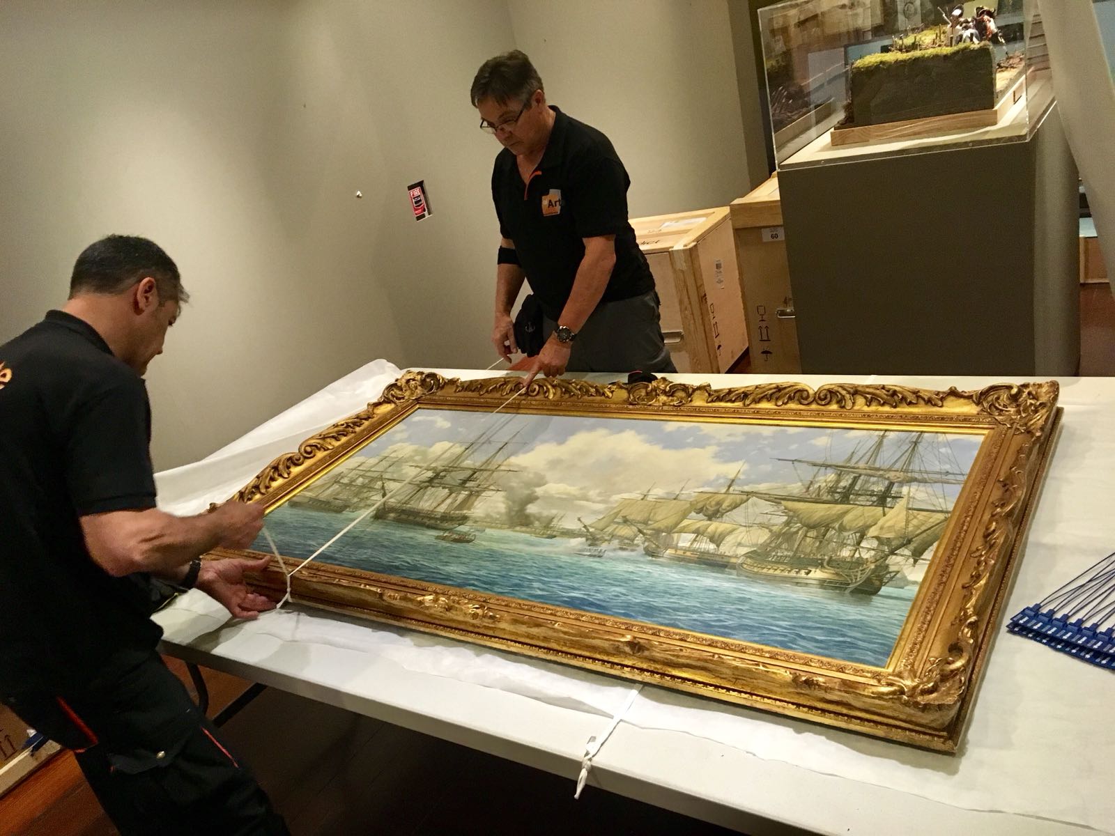 Dos técnicos de montaje miden y preparan el óleo de Pensacola tendido sobre una mesa cubierta, con cajas de embalaje y un diorama al fondo