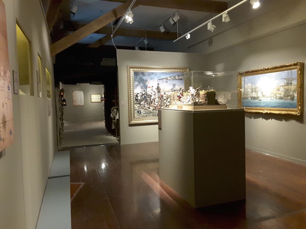 Sala de museo con suelo de madera, dos óleos enmarcados en dorado a ambos lados y una vitrina central con un diorama de soldados