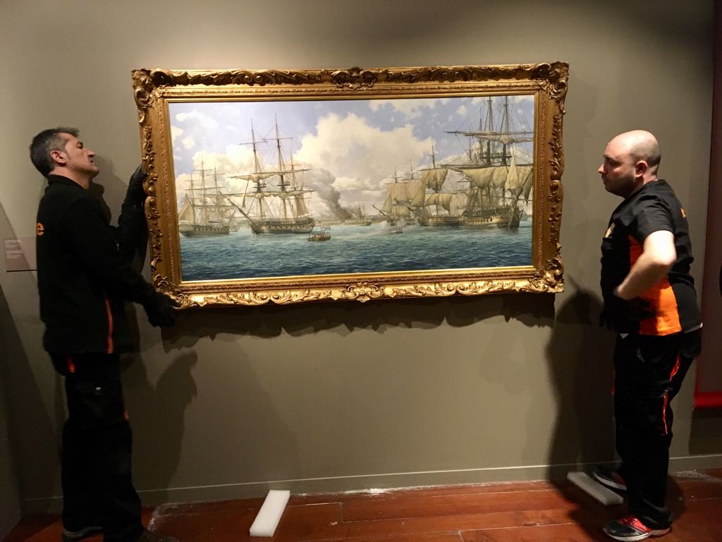 Dos técnicos vestidos de negro y naranja sostienen y ajustan el óleo de Pensacola en su marco dorado sobre una pared clara