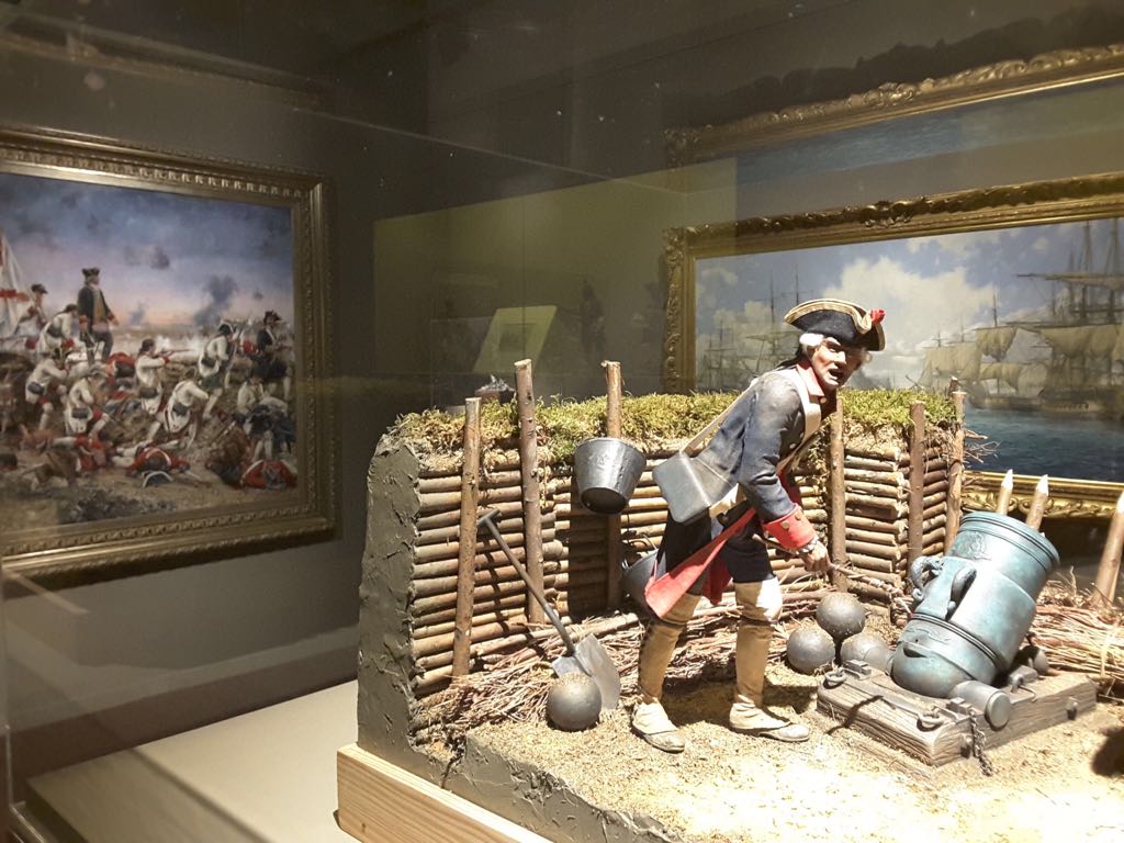 Diorama en vitrina con un artillero de casaca azul junto a un mortero de bronce, con el óleo de Pensacola enmarcado al fondo